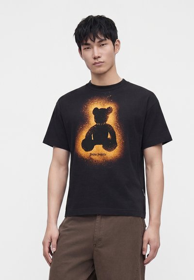 Νέος άνδρας που φοράει μαύρο T-shirt με πορτοκαλί γραφικό αρκούδας σε σπρέι και καφέ παντελόνι, στέκεται μπροστά σε απλό λευκό φόντο.