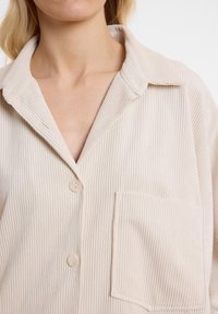 Cremfarbens Cordhemd mit Notch-Kragen, Knopfverschluss und einer Brusttasche. Der Stoff weist eine vertikal gerippte Textur auf.
