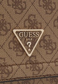 Borsa in materiale sintetico marrone con motivo a fantasie, presenta un logo triangolare dorato con la scritta "GUESS USA" e un design a punto interrogativo. Dettaglio della superficie testurizzata.