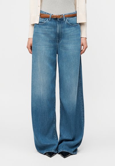 Femme portant un jean large bleu clair avec une ceinture marron, un cardigan beige, un haut gris et des chaussures noires à bout pointu.