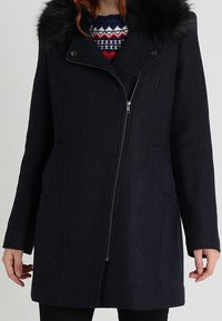 Kvinna iklädd en mörk marinblå kappa med en asymmetrisk dragkedja och krage av svart fuskpäls över en mönstrad tröja.