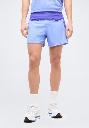 Osoba ubrana w jasnofioletowe sportowe spodenki, białe skarpetki i białe buty do biegania, stojąca na tle jednolitego tła.