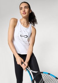 Top sportivo bianco con grafica circolare nera e simbolo dell'infinito, abbinato a leggings neri. Il modello tiene in mano una racchetta da tennis.