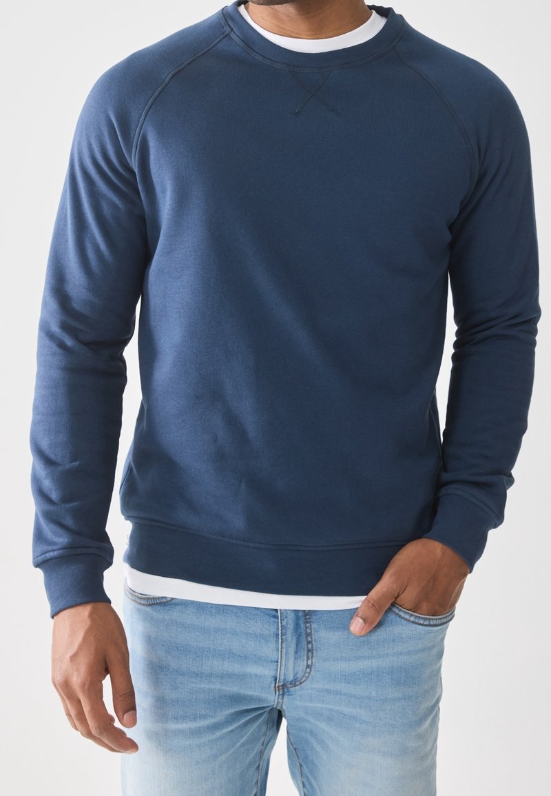Homme portant un sweat-shirt bleu marine à manches longues par-dessus une chemise blanche, avec un jean bleu clair, debout avec une main dans la poche.
