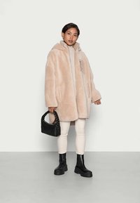 Femme portant un manteau en fausse fourrure beige oversize, un col roulé crème, un pantalon crème, des bottes noires épaisses, tenant un sac à main tressé noir, debout sur un sol gris.