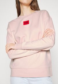Personne portant un sweat-shirt rose clair avec un écusson avec le logo rouge « HUGO », bras croisés, montrant le torse supérieur et une partie d’un jean bleu clair.