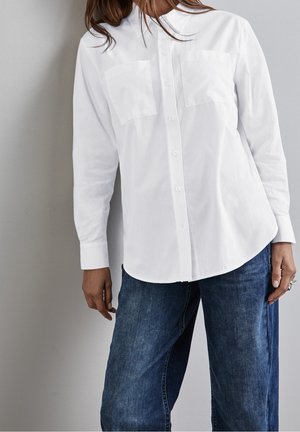 Femme portant une chemise blanche à manches longues avec deux poches poitrine et un jean en denim bleu, debout devant un mur clair uni.