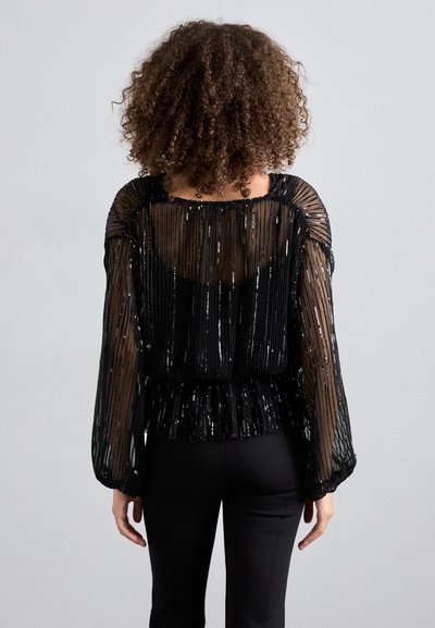Haut noir transparent avec un tissu plissé et des détails métalliques, avec des manches longues et un ourlet peplum, porté avec un pantalon noir ajusté.