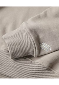 Primo piano di un tessuto color beige chiaro per felpa, con polsini a coste e un logo ricamato in bianco, che enfatizza morbidezza e texture.