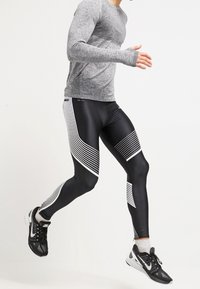 Mannen sportoutfit met een grijs langemouwtop en zwarte leggings met witte diagonale strepen; de stof lijkt rekbaar en gestructureerd.