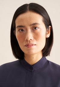Camicia button-up blu scuro con colletto a banda, texture liscia e riflesso sottile; il modello presenta capelli neri corti e trucco minimale.