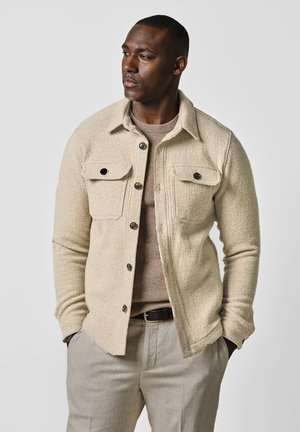 Giacca beige con texture, dotata di bottoni sul davanti, due tasche sul petto e colletto, indossata sopra un maglione beige chiaro e pantaloni beige.