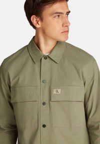 Chaqueta verde oliva con diseño de cuello, dos bolsillos frontales, botones a presión negros y una etiqueta con logo en el pecho. Textura suave.