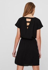 Schwarzes Kleid mit kurzen Ärmeln, elastischem Bund und einem Rückenausschnitt, der drei vertikale Riemen zeigt. Leichter, fließender Stoff.