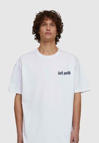 Fehér pamut póló, laza fazonnal, kerek nyakkal, rövid ujjakkal, a bal oldalon szürke "elveszett ifjú" felirattal. Simaszövet.