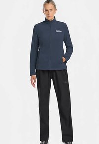 Marineblauw fleecejack met een ritssluiting aan de voorkant en een logo; gecombineerd met zwarte, loszittende broek; voorzien van een hoge kraag en zakken.