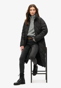 Sort quiltet lang frakke over en grå sweater, parret med sorte jeans og snørelædersko, siddende på en skammel mod en hvid baggrund.