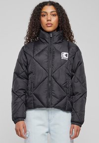 Juodas pūkinis švarkas su aukštu apykaklė, deimanto formos siuvimo modeliu, užtrauktuku ir logotipu kairėje krūtinės pusėje. Yra elastiniai rankogaliai ir apatinė dalis.