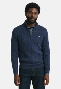 Dunkelblauer Kabelstrickpullover mit hohem Kragen und Reißverschluss. Mit gerippten Bündchen und Saum, getragen über einem hellen Jeanshemd.