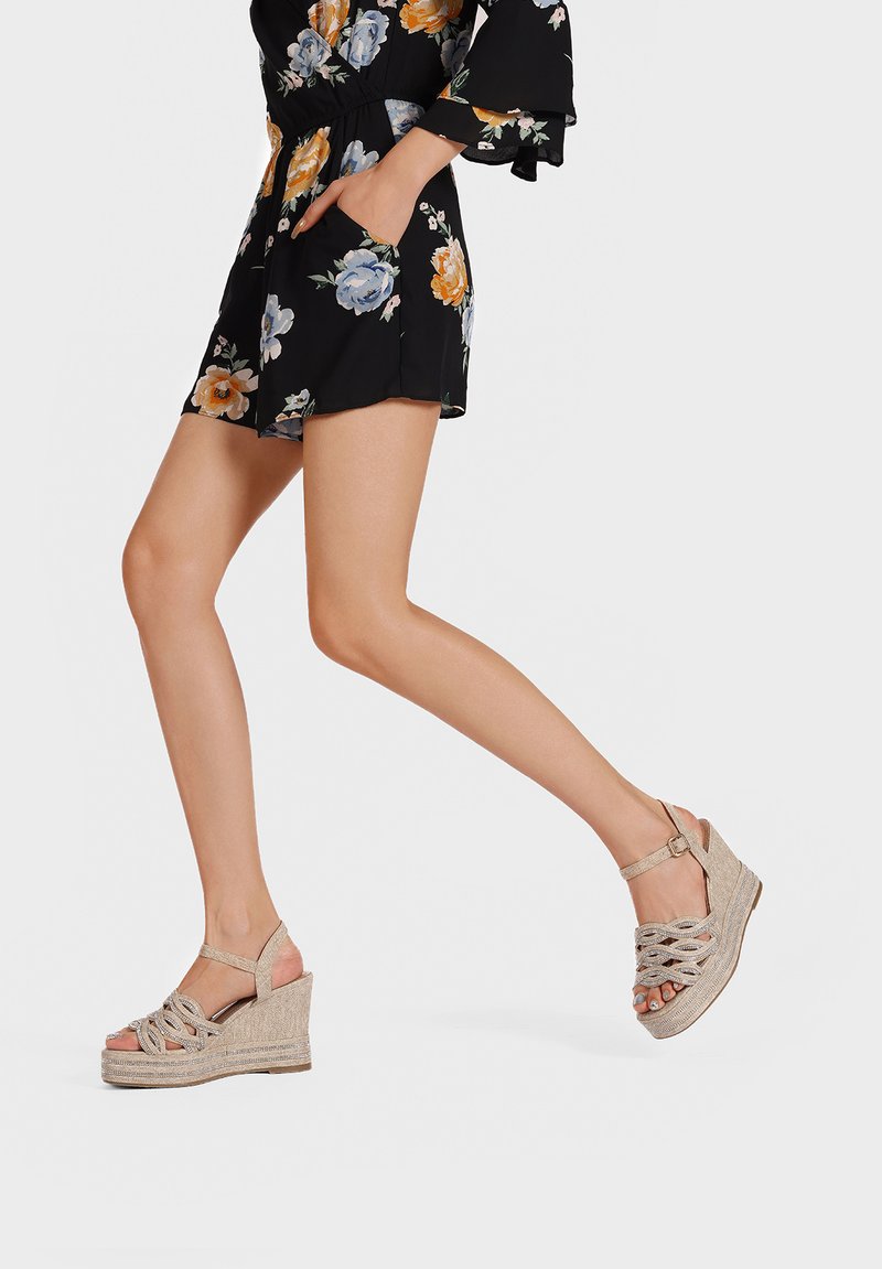 Sandali a zeppa beige con cinturini intrecciati, tacco avvolto e cinghietta alla caviglia. La modella indossa un playsuit nero a fiori mentre cammina.