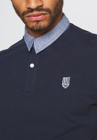Marinblå polotröja med en chambray-krage, utrustad med en vit knapp och en liten broderad sköldlogotyp på bröstet.