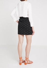 Falda negra con lunares blancos, de cintura alta y cierre en la parte trasera. Combinada con una camisa blanca de manga larga y sandalias naranjas con tiras.