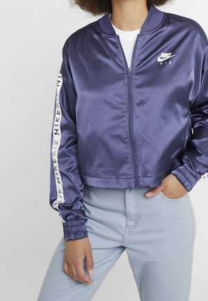 Personne portant une veste zippée en satin Nike Air violette avec le logo sur la poitrine et les manches, associée à un jean bleu clair.