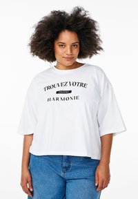 White cotton t-shirt with short sleeves, featuring black text "TROUVEZ VOTRE PROPRE HARMONIE" in a bold, modern font. Cropped design.