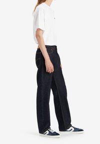 Personne portant un t-shirt blanc, un jean large foncé et des baskets bleu marine, debout de profil sur un fond blanc.