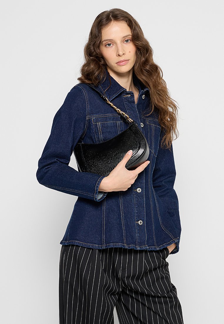 Jeune femme avec de longs cheveux ondulés portant une veste en denim foncé, un pantalon à rayures noires et blanches, tenant un sac à main noir texturé avec une bandoulière en chaîne.