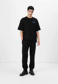 Camiseta de algodón negra con mangas cortas y logo azul, combinada con pantalones de chándal negros con un acento azul, complementada con zapatos negros.