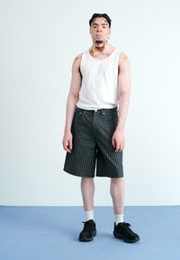 Valge tank top, rohelise ja musta ruuduliste põlvekeksu lühikesed püksid, valged lühikesed sokid, mustad spordijalatsid, seismas helesinisel põrandal.