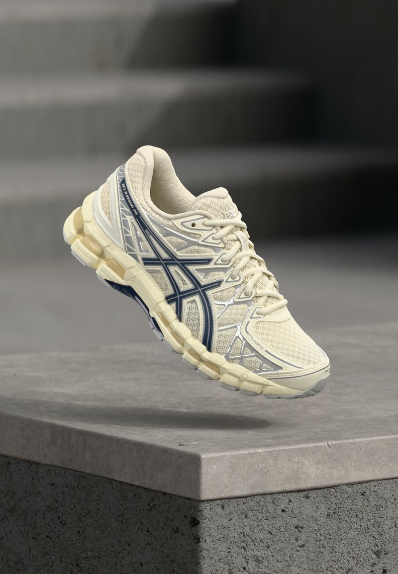 Κρέμα και ναυτικό μπλε παπούτσι τρεξίματος ASICS κρεμασμένο στον αέρα πάνω από μια σκυρόδεμα ledge με θολές σκάλες στο φόντο.