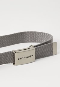 Pelēka auduma josta ar teksturētu apdari, kurai ir metāla sprādze ar iegravētu uzrakstu "carhartt". Vienkāršs dizains ar plakaniem maliņām.
