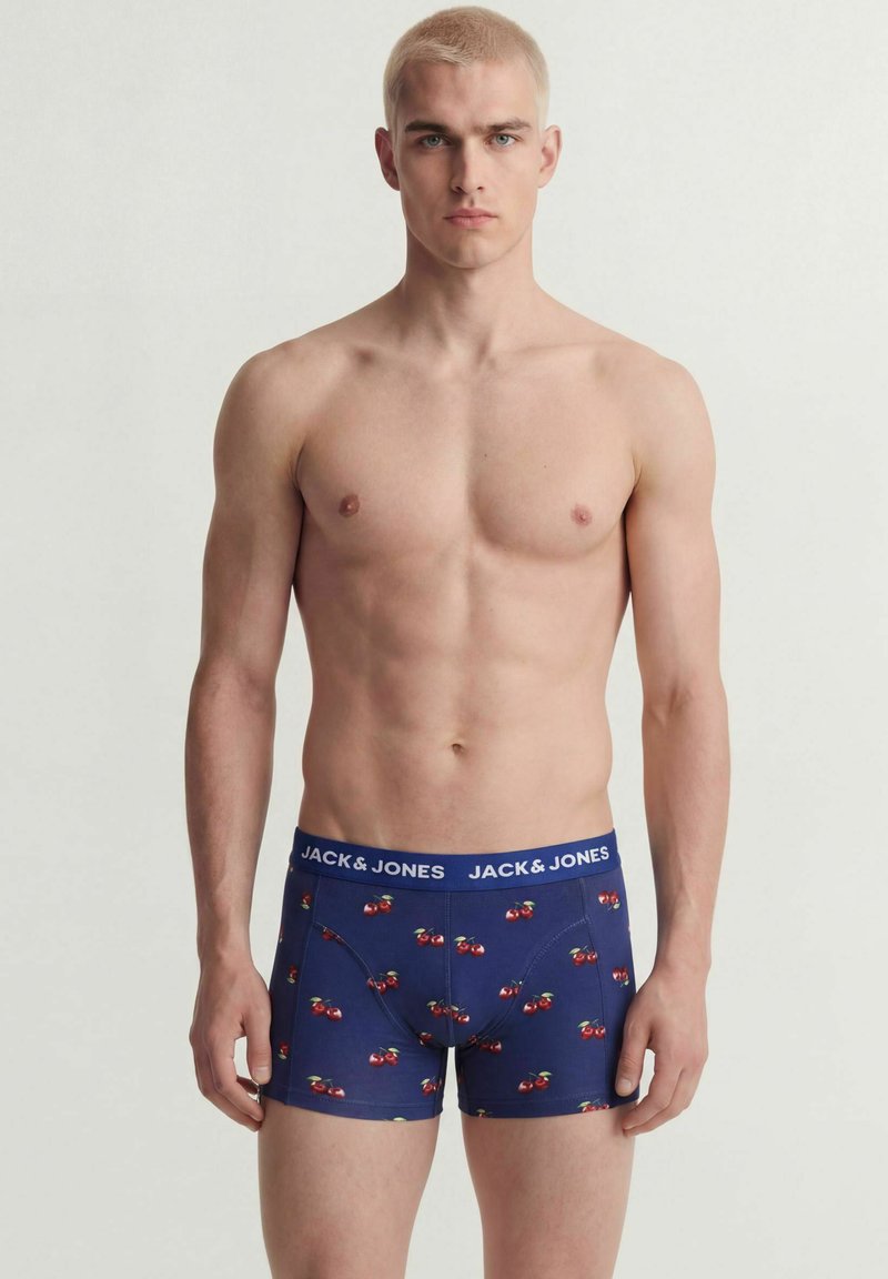 Jeune homme aux cheveux blonds courts portant un boxer bleu marine avec un motif de cerises et le logo Jack & Jones sur la ceinture, debout sur un fond blanc.