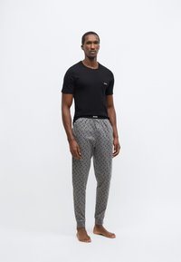 Maglietta nera in cotone con un piccolo logo, abbinata a pantaloni della tuta grigi con un motivo sottile e polsini elastici, modello in piedi a piedi nudi.