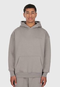 Giovane uomo che indossa un hoodie grigio con tasca frontale e pantaloni della tuta grigi abbinati, in piedi di fronte a uno sfondo chiaro e semplice.