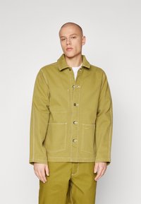 Chaqueta de algodón verde con cierre frontal de botones, dos bolsillos en el pecho y detalles de costura blanca. Lleva puesta sobre una camisa blanca, combinada con pantalones a juego.