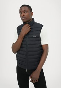 Schwarzer puffer Weste mit hohem Kragen, gestepptem Design, vertikalen Nähten und Reißverschlusstaschen. Verfügt über ein kleines Logo auf der Brust.