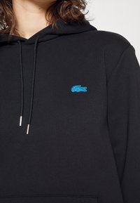 Sudadera con capucha negra de tejido suave, con bolsillo tipo canguro y capucha con cordón. Destaca el logo azul bordado del cocodrilo en el pecho izquierdo.