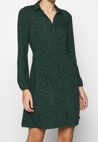 Abito verde a maniche lunghe con motivo floreale nero, con bottone frontale e colletto, indossato da una persona in piedi.