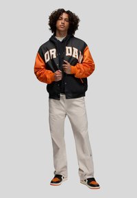 Svart och orange varsityjacka med "ORA DAN"-bokstäver, svart hoodie under, beige byxor och svarta/orange sneakers.