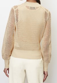 Beżowy sweter dzianinowy z siateczkowym wzorem, posiadający długie rękawy i ściągacze u mankietów. Tył jest w większości otwarty z teksturowanym wzorem.