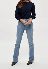 Eine Frau steht vor einem neutralen Hintergrund, trägt einen navyblauen Pullover und hellblaue Schlagjeans, kombiniert mit olivgrünen Schuhen.