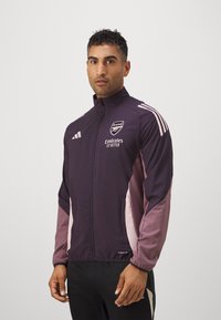 Dunkelviolette und rosa Sportjacke mit hohem Kragen, Reißverschluss vorne und weißen Adidas-Streifen; verfügt über das Arsenal-Logo und den Text 'Emirates'.