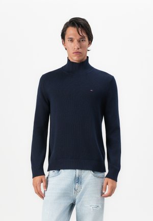 Man in een marineblauwe coltrui en lichtblauwe ripped jeans, staand tegen een simpele witte achtergrond.