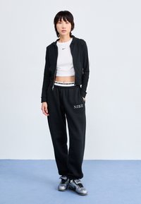Sudadera negra con cremallera y rayas blancas, camiseta blanca corta, pantalones de chándal negros con bordado 'NIKE' y zapatillas deportivas plateadas.