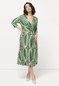 Grünes gemustertes Wickelkleid mit Dreiviertelärmeln, V-Ausschnitt, tailliertem Design und einem fließenden Rock. Enthält beigefarbene Blättermotive.