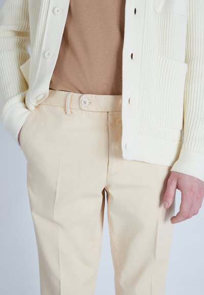 Cardigan a maglia color crema indossato sopra una camicia beige, abbinato a pantaloni beige chiaro con chiusura a bottone e una texture liscia.