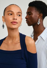 Jūros laivyno rausvas vir�šus su off-shoulder dizainu, derinamas su sidabriniais abstrakčiais formos auskarais. Fone matoma modelis su dryžuotu marškiniu.